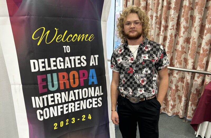 Jake Hinds poses with a board that says he is at the Delegates at Europa International Conference.