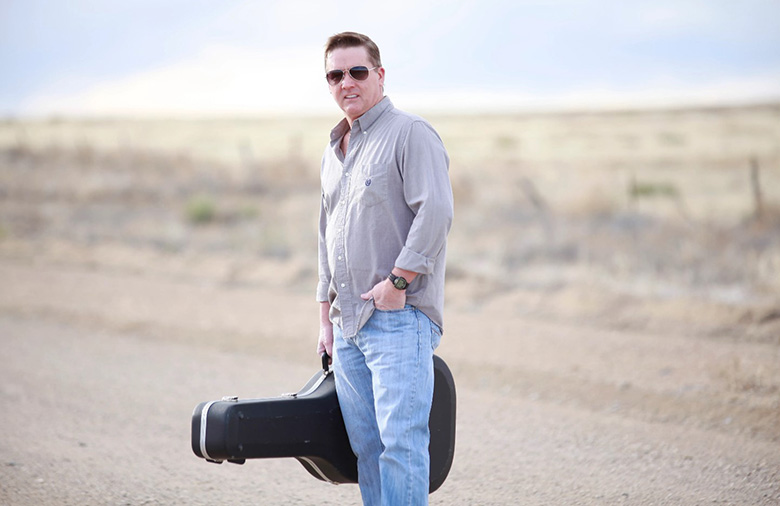 Lex Nichols holding guitar case on a dirt road.