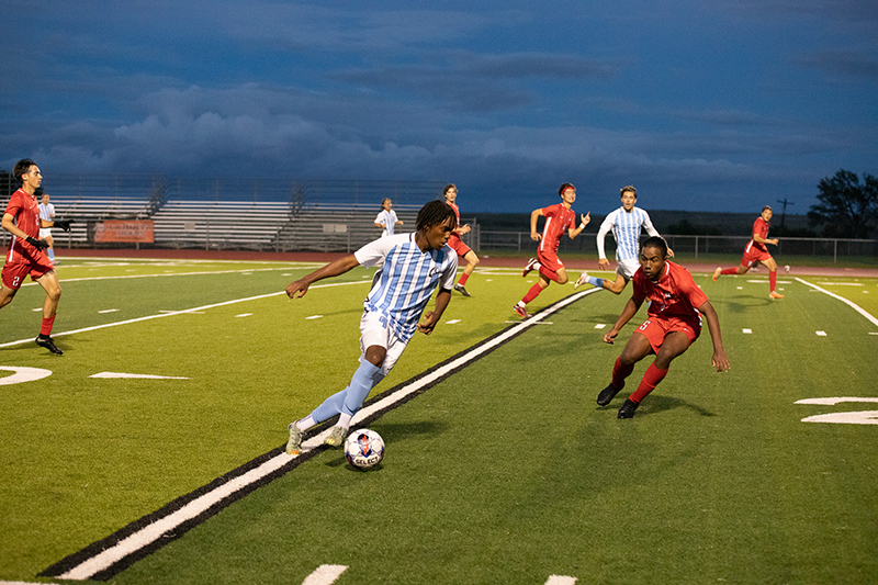 Soccer player running down a field with the ball with bleachers and clouds in the background.