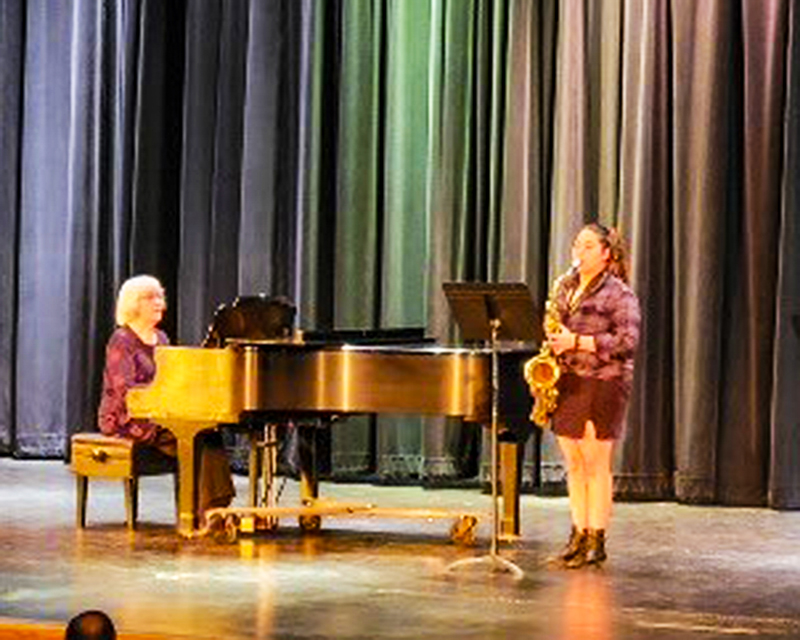 A student playing a saxophone on a stage with a woman playing the piano accompanying her.