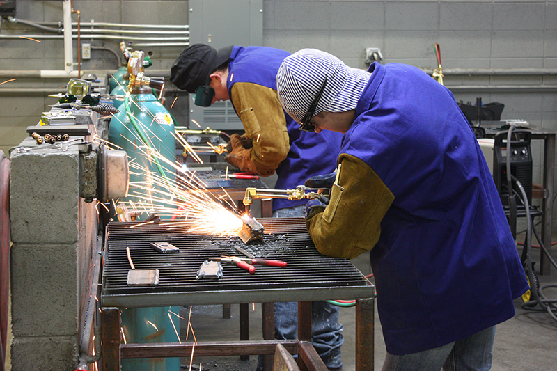 Welding students leaning over a table focused on their welding project with sparks flying.